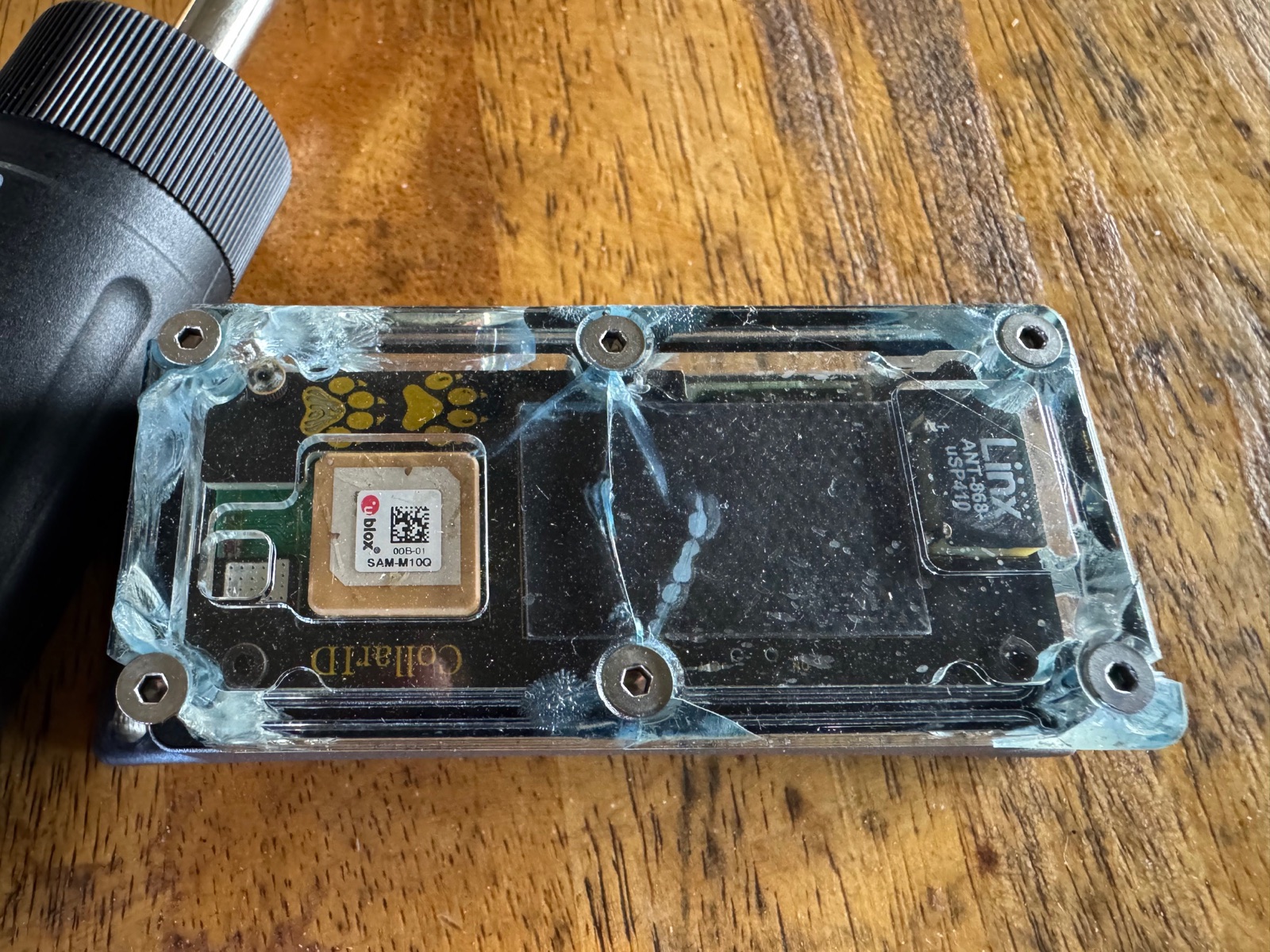 A CollarID cover with cracks running out from every screw hole, on a wooden table next to the torque driver
